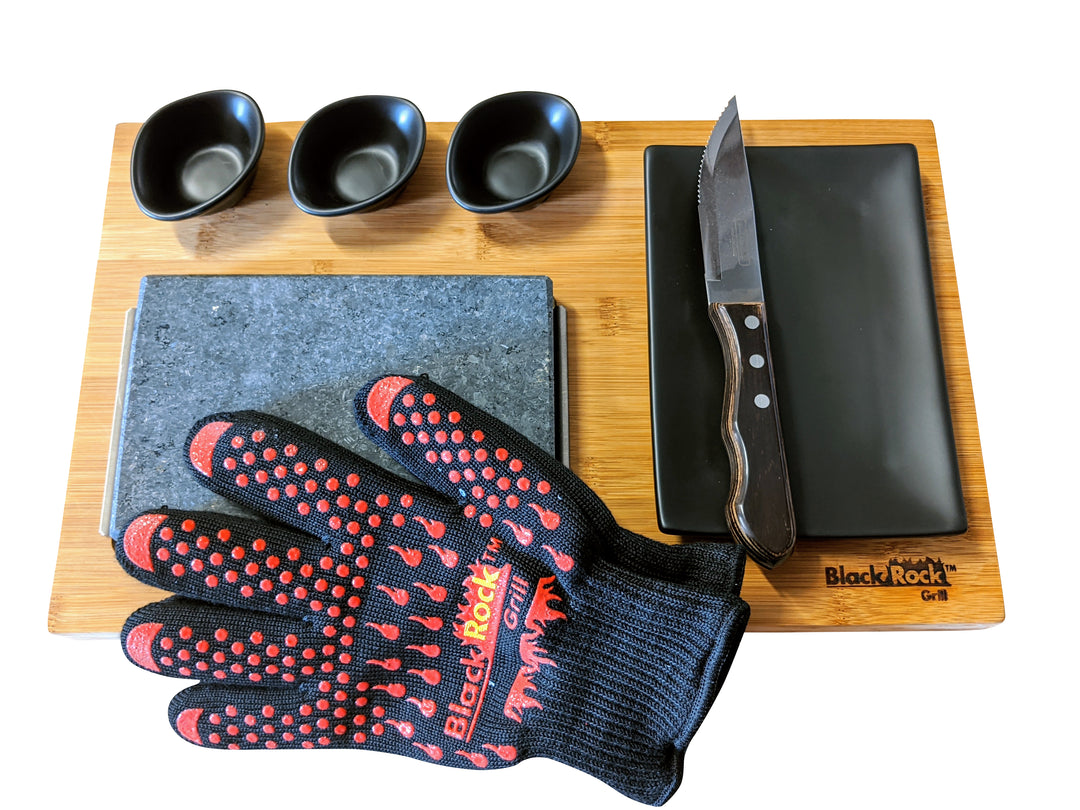 The Black Rock Grill Steak Stones Cooking Gift Set includes a bamboo serving board with a knife, a black rectangular plate, three small bowls, a granite slab for hot stone cooking, and a heat-resistant glove featuring red text and dots for safely handling Black Rock Grill steak stones.