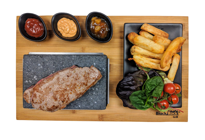 The Black Rock Grill Matt Black Steak Stones Cooking Set offers a sizzling steak experience on a hot stone with three dipping sauces, crispy fries, and a small salad with lettuce and cherry tomatoes, all beautifully presented on an artfully arranged wooden board by Black Rock Grill.