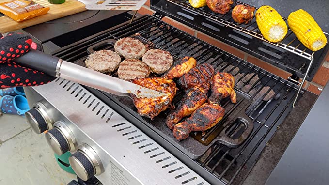A hand in a red and black glove uses tongs to turn chicken on the Black Rock Grill Large Reversible Cast Iron Griddle Pan (46x23cm) with silicone grip handles as burgers and corn cook nearby; grill knobs and a cutting board are visible.