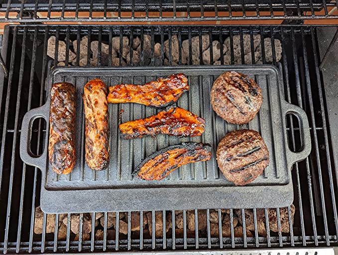 On the Black Rock Grill Large Reversible Cast Iron Griddle Pan (46x23cm) with silicone grip handles, sausages, bacon, and burger patties sizzle over charcoal, displaying grill marks that seal in classic BBQ flavor. Includes marinating oil brush.
