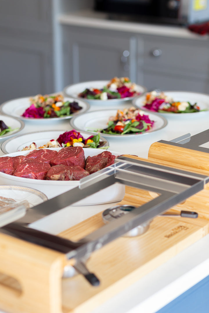 A dining setup includes white salad plates with colorful veggies and a tray of raw red meat beside the Black Rock Grill Sharing Steak Stones Grill Gift Set. A kitchen counter and cabinets in the background provide a perfect setting for sharing hot stone cooking experiences.
