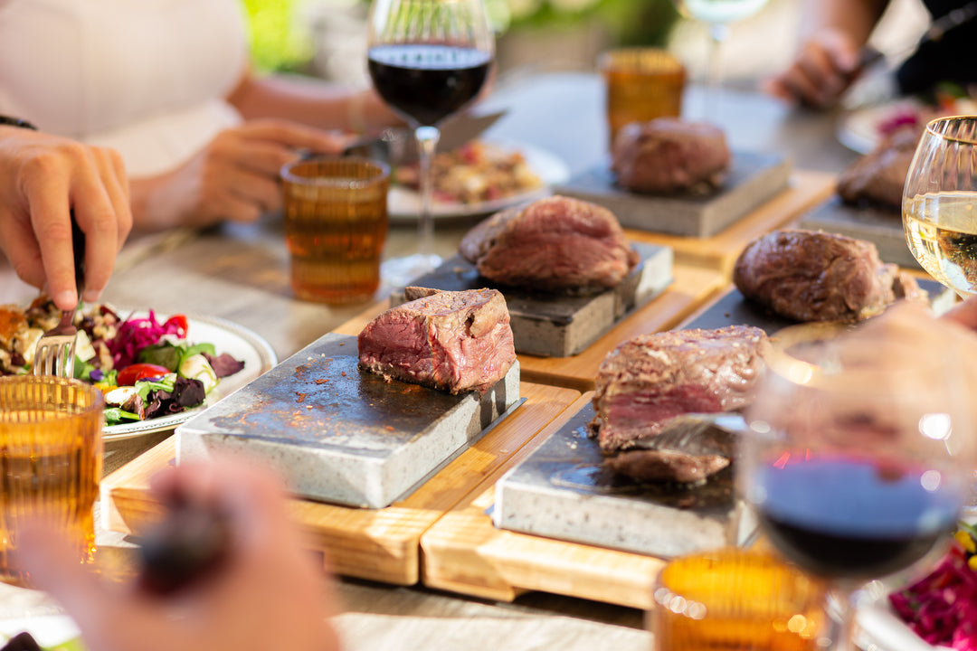 The Black Rock Grill Steak Stone Cooking Set transforms the dining experience with sizzling hot stones and steaks, complemented by red and white wine. Colorful salads in bowls encourage healthy eating, while hands with utensils signify an active meal under a bright, sunlit ambiance.