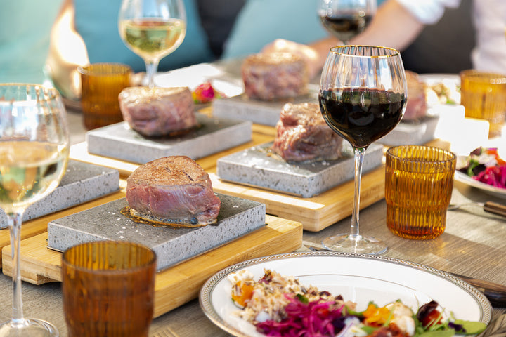 A Black Rock Grill Steak Stone Cooking Set invites the art of hot stone cooking with individual slabs for raw steaks, complemented by glasses of red and white wine, and vibrant salads that enhance a sophisticated dining environment.