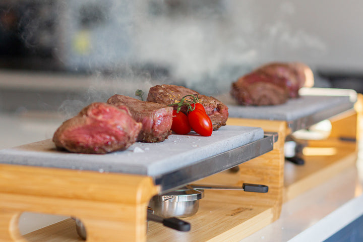 Cooked meat slices sit on the Black Rock Grill Sharing Steak Stones Grill Gift Set, garnished with cherry tomatoes. The setup includes a wooden stand and a heating element underneath, releasing steam. This Black Rock Grill cooking experience is ideal for sharing, set against a blurred background.