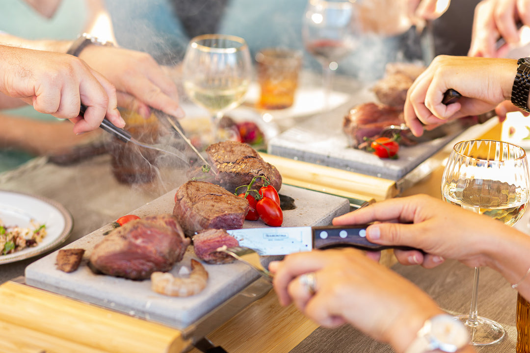 People gathered around a table, slicing cooked steaks on Black Rock Grill Sharing Steak Stones Grill Gift Set platters. Glasses of wine and small tomatoes are present. Steam rises from the food, and hands with utensils show shared enjoyment of this unique hot stone cooking experience by Black Rock Grill.