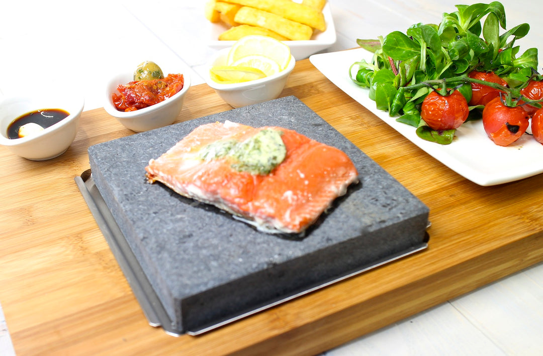 The Black Rock Grills Steak Stone Grill & Effeuno Pizza Oven Combo Package features raw salmon sizzling on a lava stone, paired with a cherry tomato salad, potato wedges, and bowls of sauce, lemon slice, sun-dried tomatoes, and an olive—all artfully presented on a wooden board.