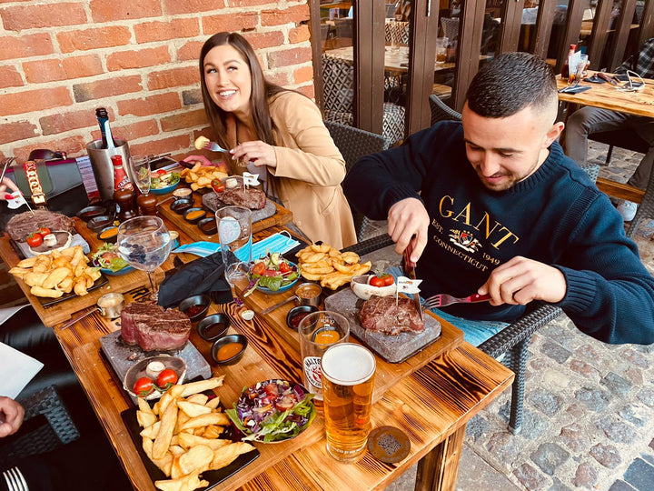 A couple enjoys an interactive meal using the ROXY12 by Black Rock Grill, with steak and fries on plates at their wooden table. The woman smiles as the man cuts his steak, enriched by condiments and drinks. Theyre set against a charming brick wall backdrop in an outdoor setting.