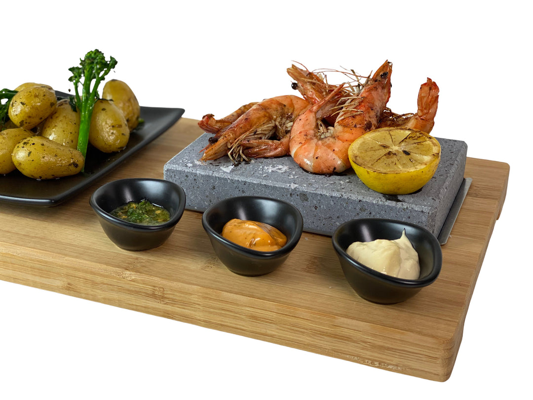 The Black Rock Grill ROXY45 set features a hot stone with sizzling grilled prawns and lemon, plus three sauce bowls. Served on a rustic board, it includes a black plate of roasted potatoes and broccolini for an interactive and healthy dining experience.
