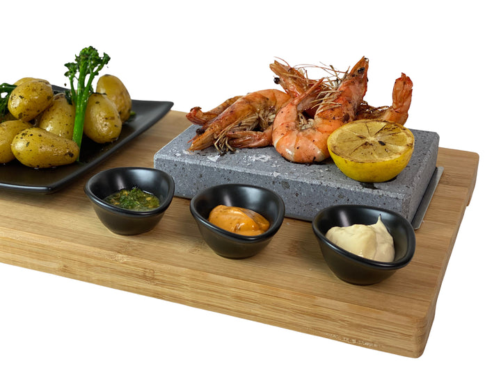 The Black Rock Grill ROXY45 set features a hot stone with sizzling grilled prawns and lemon, plus three sauce bowls. Served on a rustic board, it includes a black plate of roasted potatoes and broccolini for an interactive and healthy dining experience.