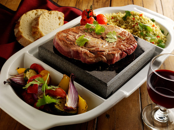 The Black Rock Grill ROXY12 set features a sizzling steak on a hot stone garnished with cilantro. Grilled vegetables, couscous, and slices of bread surround it. A glass of red wine complements the interactive dining experience on the wooden table, creating a healthy meal moment.