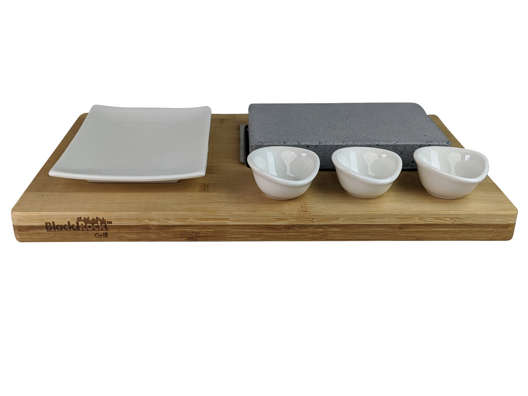 The Black Rock Grill Steak On The Stone Set includes a wooden serving board with a hot stone slab, perfect for cooking steak, three small white bowls, and a square white plate. This set invites you to embrace the unique hot stone dining experience by cooking your own meal.