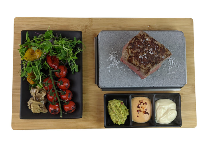 The Black Rock Grill Black Steak Stone Cooking Set by Black Rock Grill features a sizzling stone on a bamboo board for cooking steak, served with arugula, cherry tomatoes, artichokes on a black plate, and sides of guacamole, spicy sauce, and aioli—ideal for healthy dining enthusiasts.