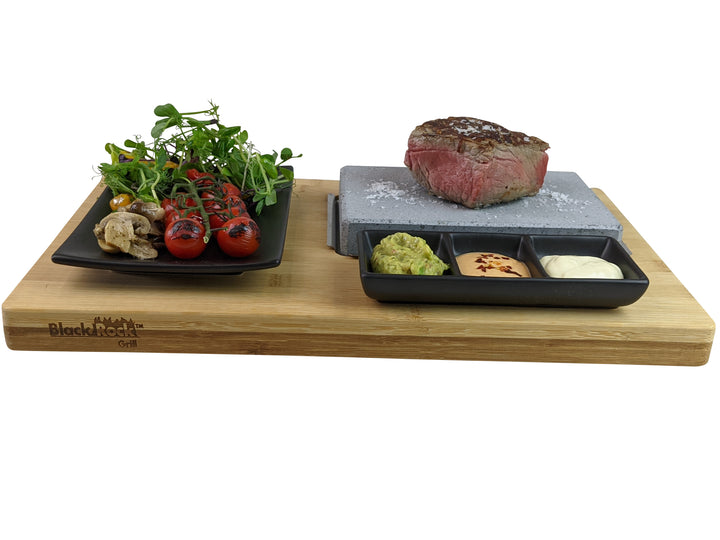 Elevate your dining experience with the Black Rock Grill Black Steak Stone Cooking Set. Enjoy a sizzling steak on a hot stone alongside grilled tomatoes, mushrooms, greens, and three sauce ramekins, beautifully arranged on a wooden board for a healthy and elegant meal.