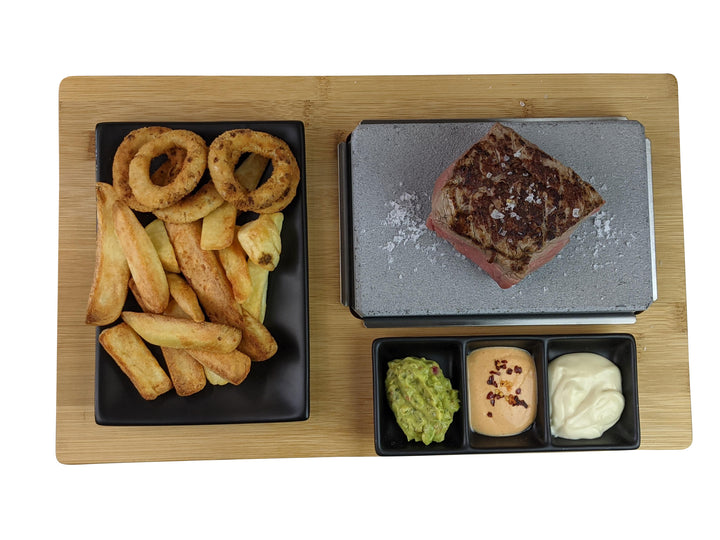 A sizzling steak from the Black Rock Grill Black Steak Stone Cooking Set is displayed on a wooden board with thick-cut fries and onion rings. Three small black dishes offer guacamole, creamy orange sauce, and mayonnaise for the perfect hot stone cooking experience.