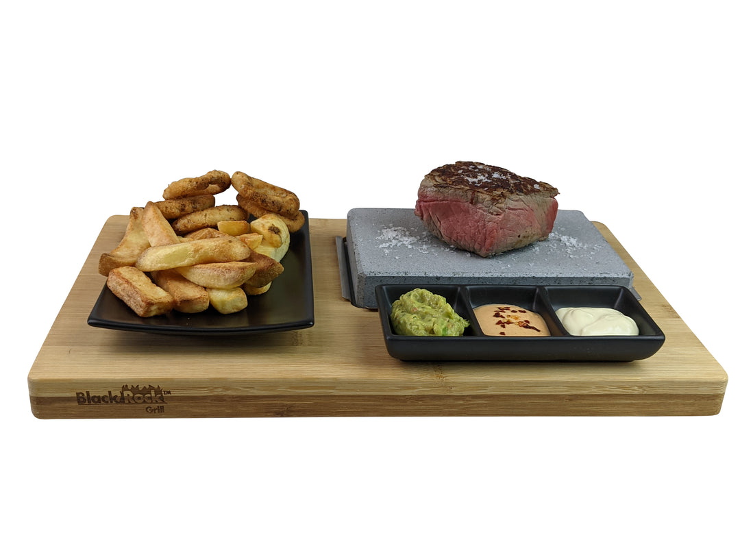 The Black Rock Grill Black Steak Stone Cooking Set presents a grilled steak on a hot stone, accompanied by guacamole, reddish sauce, and mayonnaise. Adjacent is a black dish with crispy potato wedges and onion rings, highlighting Black Rock Grills signature healthy dining approach.