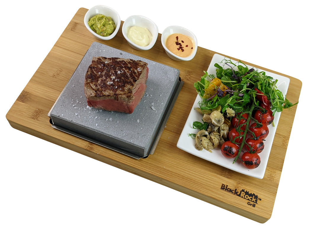 A Black Rock Grill board features a steak sizzling on a volcanic lava stone, accompanied by bowls of guacamole, mayonnaise, and spicy sauce. A side plate holds grilled vegetables and cherry tomatoes. This display aligns with the Steak Stone Grill & Effeuno Pizza Oven Combo Package branding.