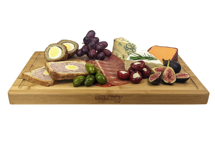 The Black Rock Grill Large Wooden Serving Board is ideal for hosting, showcasing various snacks like pâté with eggs, Scotch eggs, green olives, grapes, red-stuffed peppers, salami, halved figs, and blue and orange cheeses. Enjoy custom laser engraving options for a personalized touch.