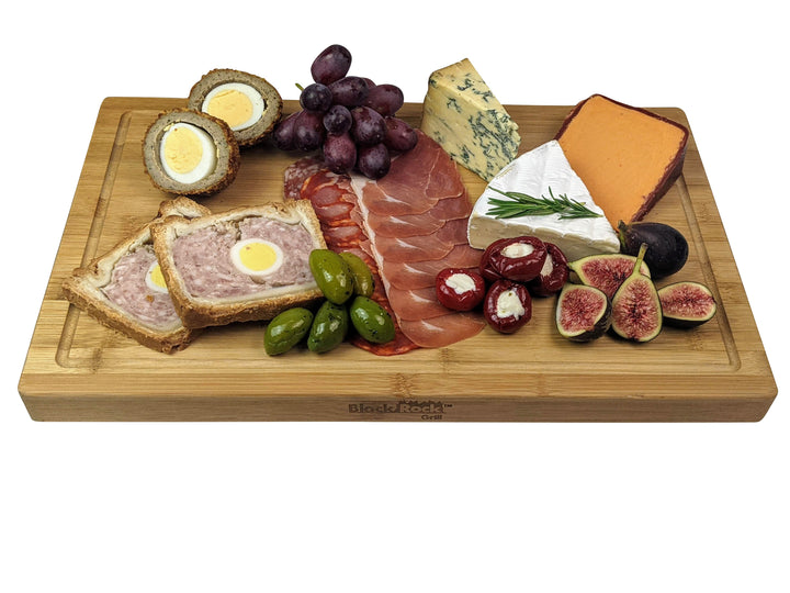 The Large Wooden Serving Board by Black Rock Grill elegantly presents an array of foods, including scotch eggs, grapes, blue and orange cheeses, figs, olives, meat slices, stuffed peppers, and two types of pâté. This appetizing display is ideal for any gathering.