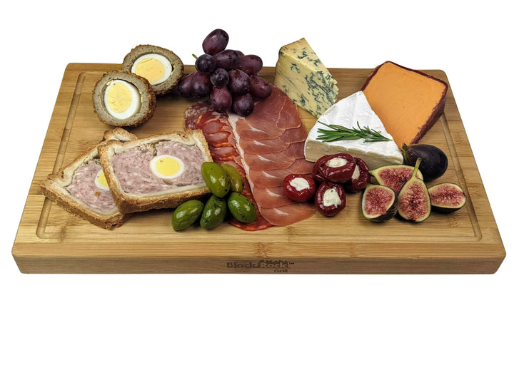 The Black Rock Grill Large Wooden Serving Board, measuring 44cm x 27cm x 3cm, beautifully presents Scotch eggs, grapes, blue and orange cheese, charcuterie, fig slices, stuffed peppers, pâté with an egg slice, and green olives for any gathering.