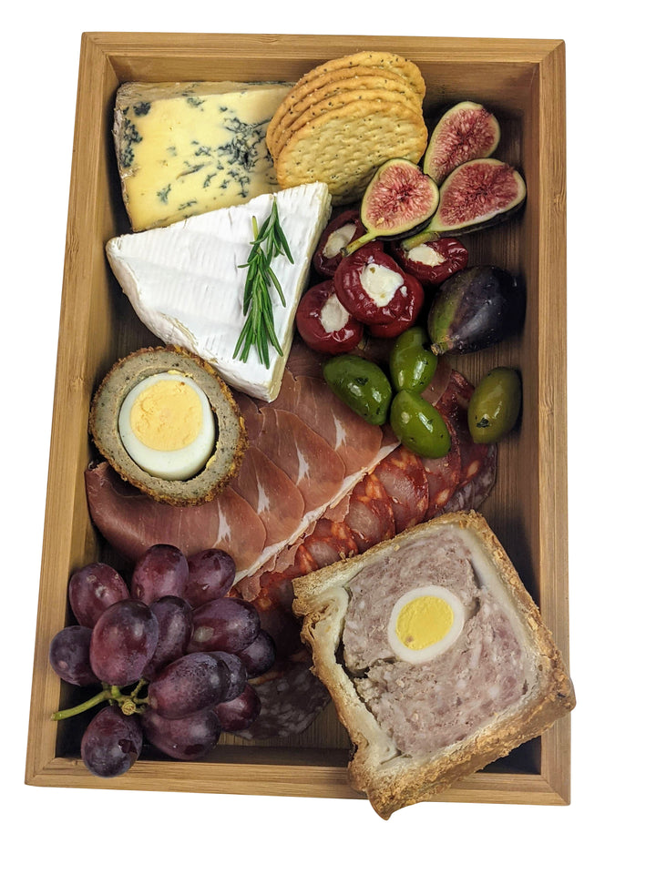 The Black Rock Grill Bamboo Wooden food presentation tray elegantly showcases assorted cheeses, crackers, figs, olives, stuffed peppers, sliced meats like prosciutto, Scotch egg, and pork pie with egg, alongside a bunch of purple grapes for an exquisite display.