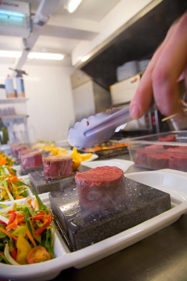 Using the ROXY12 set by Black Rock Grill, a hand with tongs lays a raw steak on a hot stone, steam rising for an interactive experience. In the foreground are white trays with fresh vegetables and colorful salads, creating an inviting scene of healthy dining in a kitchen setting.