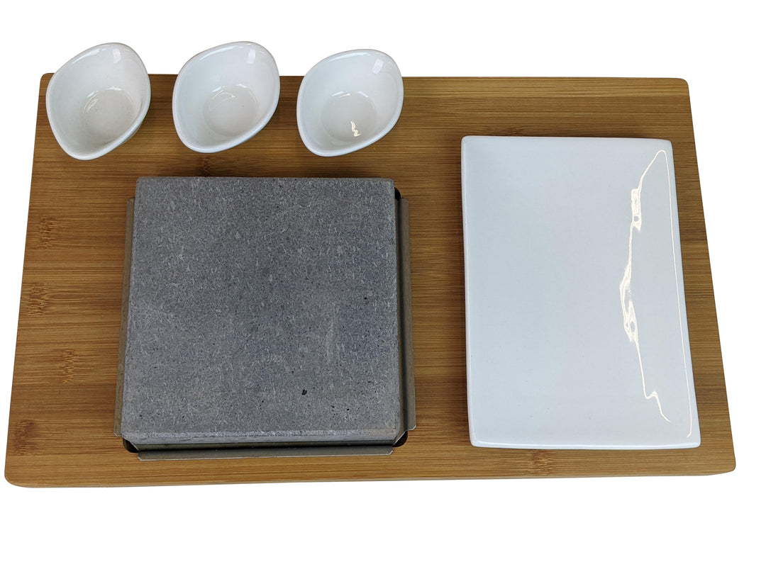 The Black Rock Grill Steak On The Stone Set includes a rectangular bamboo tray with three small white bowls, a large plate, and a gray stone slab in a square holder for hot stone dining, enabling diners to personalize their steak experience.