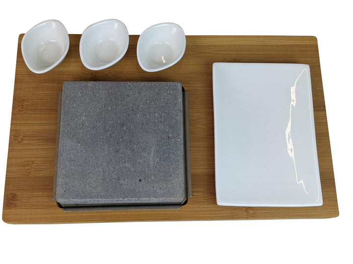 The Black Rock Grill Steak On The Stone Set includes a rectangular bamboo tray with three small white bowls, a large plate, and a gray stone slab in a square holder for hot stone dining, enabling diners to personalize their steak experience.