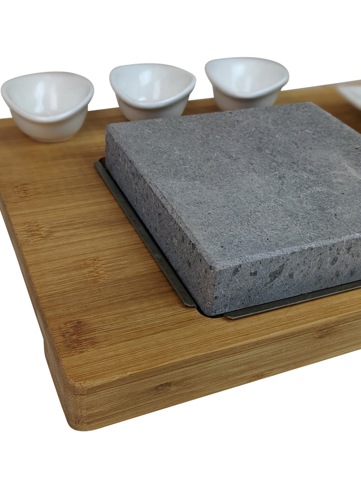 The Black Rock Grill Steak On The Stone Set offers a unique dining experience with a square stone slab on a wooden board, complete with three white bowls. Cook your meal at the table for a personalized touch.