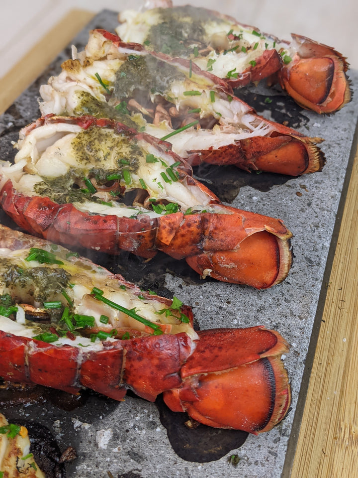 Four grilled lobster tails are beautifully presented with herb butter sauce and chives on Black Rock Grills ROXY12 set. The dish offers an interactive dining experience, letting guests enjoy table-side cooking while savoring this exquisite seafood delight.