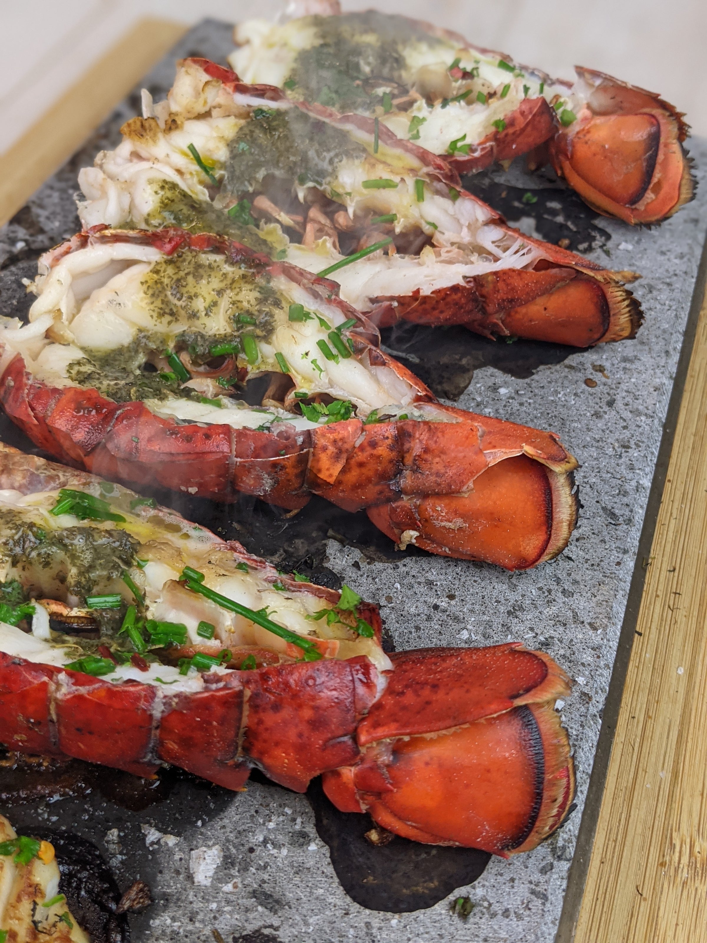 Enjoy an interactive meal with Black Rock Grills 108 Rocks set, featuring four halved lobster tails topped with herb butter sauce and chopped chives, served on a sizzling hot stone platter.