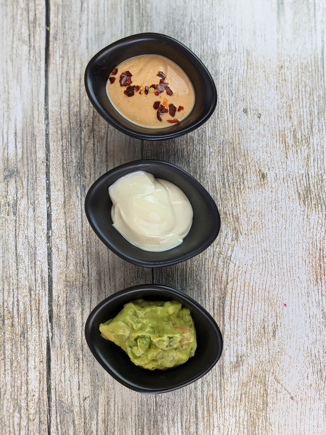 On a wooden surface, three of Black Rock Grills 12 Matt Black Porcelain Ramekin Dipping Pots hold unique sauces: the top has a spicy orange mix with red pepper flakes, the middle a creamy white delight, and the bottom fresh green guacamole. Perfect for any occasion.