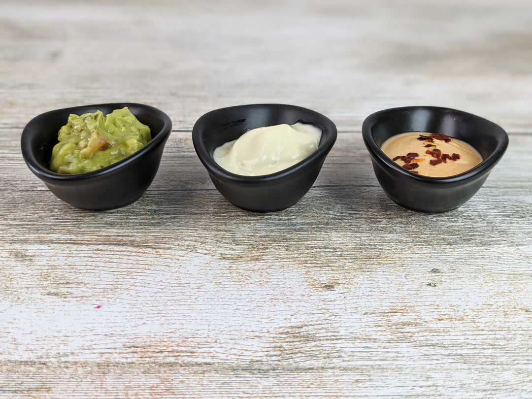 Three small ramekins from the Black Rock Grill 12 Matt Black Porcelain Ramekin Dipping Pots set rest on a wooden surface, holding guacamole, creamy white sour cream, and a tan sauce garnished with red pepper flakes.