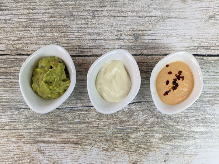 On a wooden surface, three of Black Rock Grills 12 White Porcelain Ramekin Dipping Pots hold different sauces: guacamole on the left, a creamy white sauce in the middle, and a creamy orange sauce with chili flakes on the right. Theyre also dishwasher proof.