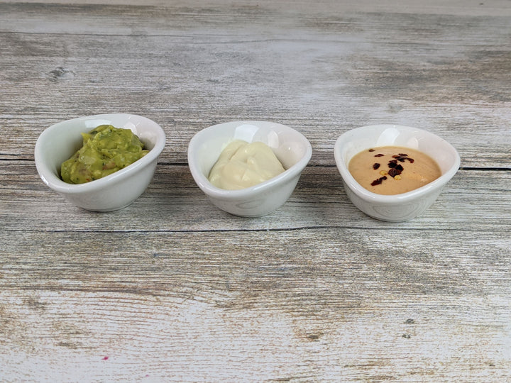 Three small Black Rock Grill White Porcelain Ramekin Dipping Pots sit on a wooden surface: guacamole on the left, mayonnaise in the middle, and spicy sauce with red pepper flakes to the right. Stylish and dishwasher proof for easy cleaning.