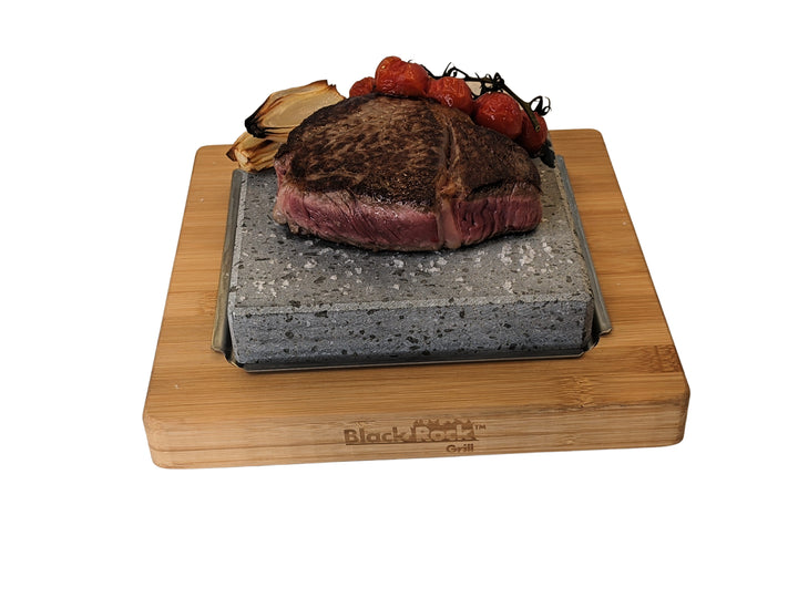A succulent steak rests on a hot Lava Stone from the NEW Black Rock Grill Lava Stone Steak Multi Pack, topped with roasted cherry tomatoes and onions on a bamboo platter. The stone sits in an engraved shallow dish by Black Rock Grill.