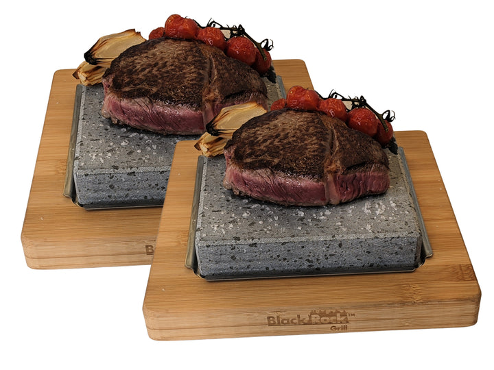 Two beef steaks are served on sizzling lava stones atop elegant bamboo platters, each with roasted cherry tomatoes and onions, ideal for hot stone cooking. The slabs are branded as the NEW Black Rock Grill Lava Stone Steak Multi Pack from Black Rock Grill.