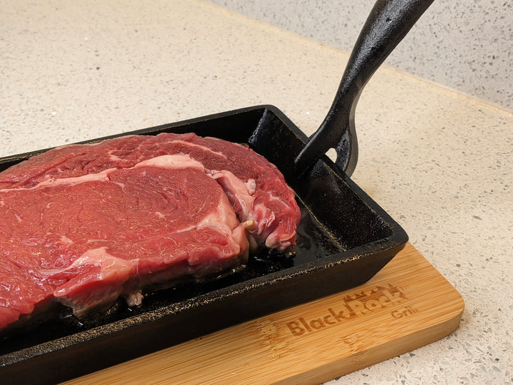 A premium quality raw steak rests on a sizzling hot Black Rock Grill Cast Iron Fajita Skillet Pan with Wood Base against a textured countertop backdrop.
