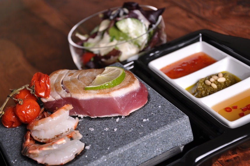 Indulge in a gourmet meal with seared tuna and shrimp on the Black Rock Grill ROXY24. Its garnished with roasted cherry tomatoes and lime slices, complemented by a side salad and three small sauce dishes, all elegantly set on a dark wooden table.