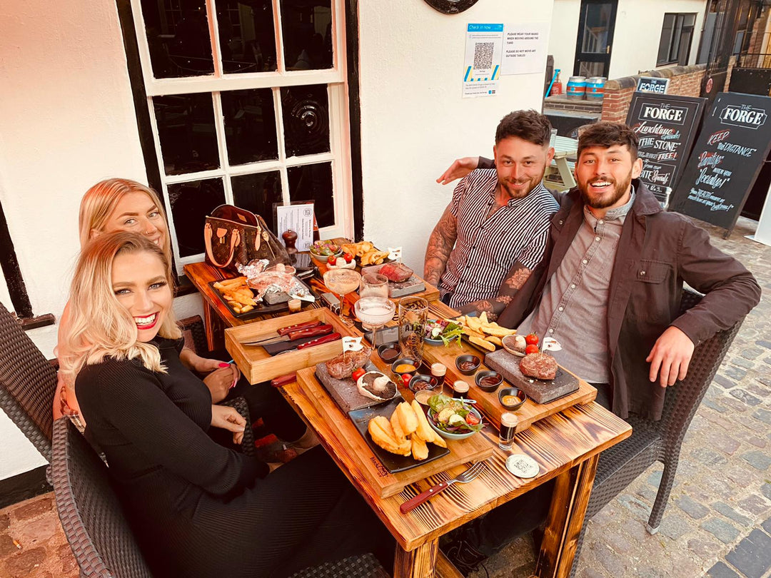 Two women and two men are seated around a wooden table at an outdoor restaurant, enjoying a delightful interactive experience with the Black Rock Grills ROXY12. The cobblestone flooring adds casual charm to this healthy dining scene as they smile over food and drinks.