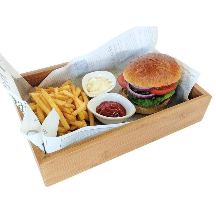 A Bamboo Wooden food presentation tray by Black Rock Grill holds a burger with lettuce, tomato, and red onion next to fries. Two small dishes of mayo and ketchup complete the setup on a tray lined with paper resembling newspaper print.