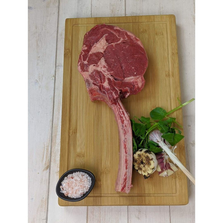 On a light wooden surface, a Black Rock Grill tomahawk steak sits on the Large Wooden Serving Board beside pink Himalayan salt in a black dish, garlic bulb, green herbs, and spring onions.