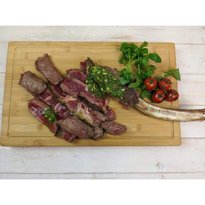 Sliced, cooked tomahawk steak rests on a sustainable Black Rock Grill Large Wooden Serving Board, topped with green chimichurri sauce. Fresh greens and vine cherry tomatoes accompany this dish, elegantly presented on the board with custom laser engraving.