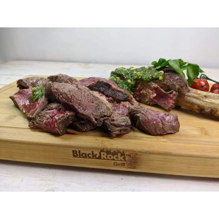 Slices of rare steak are arranged on a Large Wooden Serving Board from Black Rock Grill, with the brand logo custom laser engraved. The display is garnished with green herbs and accompanied by cherry tomatoes and arugula leaves.