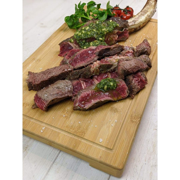 Sliced medium-rare steak garnished with green sauce on a Black Rock Grill Large Wooden Serving Board, complemented by cherry tomatoes and leafy greens.