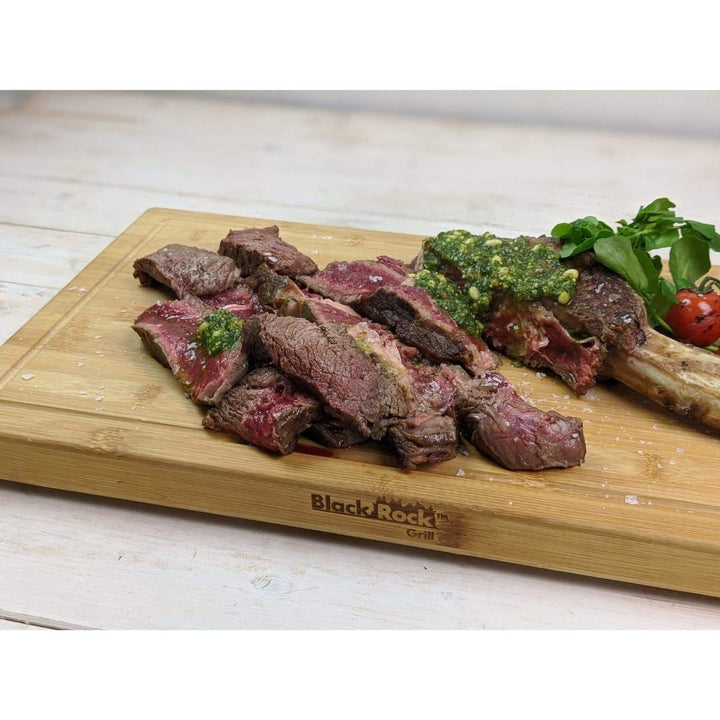 Sliced steak topped with green sauce on the Black Rock Grill Large Wooden Serving Board. A whole piece with bone is garnished with greens and cherry tomatoes. The sustainable bamboo board adds elegance to any dining setting on a light wooden surface.