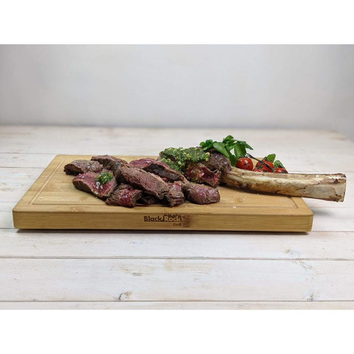 Sliced steak with green sauce sits on the Black Rock Grill Large Wooden Serving Board, alongside a bone, cherry tomatoes, and greens. This sustainable bamboo board features custom laser engraving and rests elegantly on a light wooden table.
.