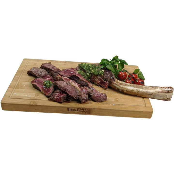 A sliced steak with green garnish rests next to a bone, accompanied by cherry tomatoes and greens on a Black Rock Grill Large Wooden Serving Board, custom laser engraved for an elegant touch.