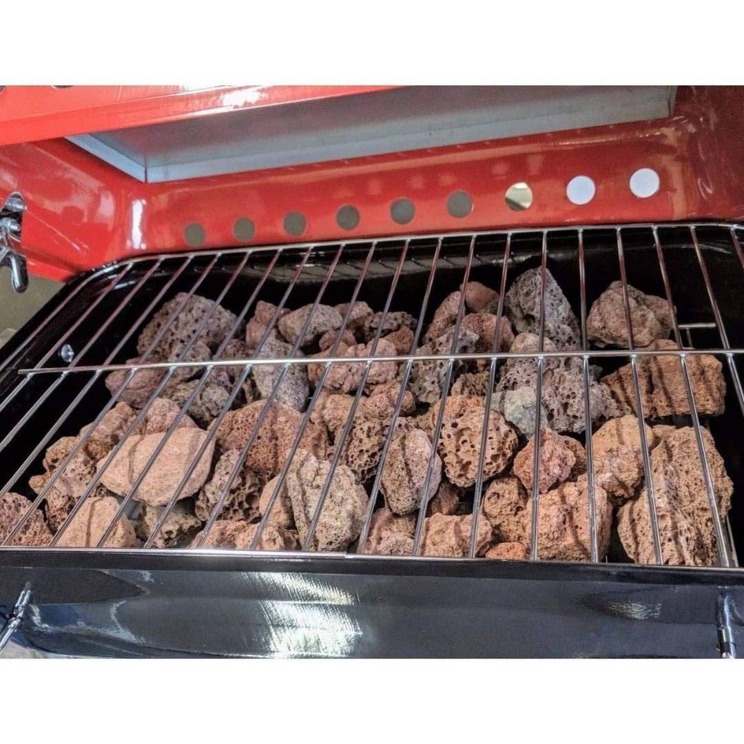 A close-up of Black Rock Grill's Lava Rocks for Grilling (4-8cm) beneath a metal grate inside an open red and black gas BBQ grill.