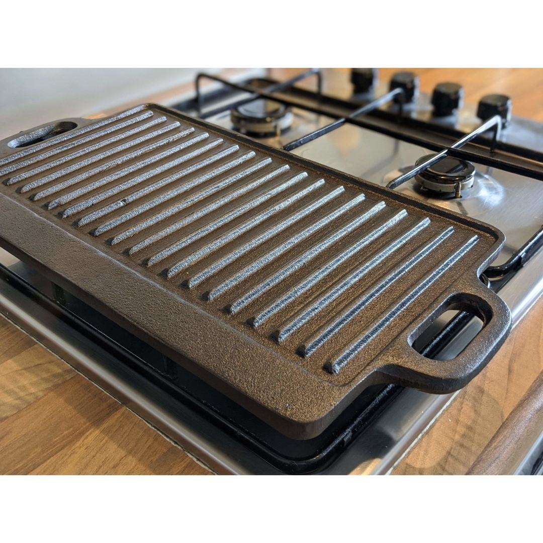 A Black Rock Grill Large Reversible Cast Iron Griddle Pan (46x23cm) with silicone grip handles sits atop a stainless steel four-burner gas stove with black knobs, with a wooden countertop in the background. Includes marinating oil brush.