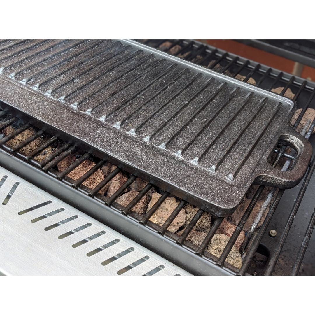 A close-up of the Black Rock Grill Large Reversible Cast Iron Griddle Pan with silicone grip handles (46cm x 23cm) on a gas grill grate, with lava rocks beneath, plus marinating oil brush included.